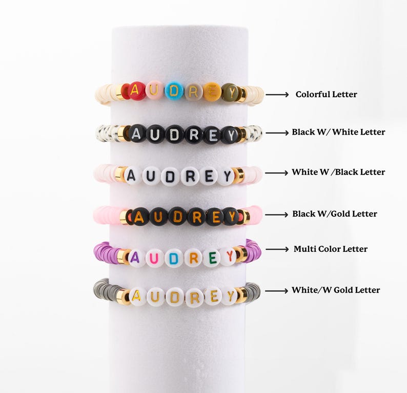 May include: Six stacked bracelets with the name "AUDREY" spelled out in colourful beads. The bracelets are made with different coloured beads and letter colours, including black, white, gold, and multi-coloured.