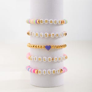 May include: Five colourful beaded bracelets with gold lettering. The bracelets spell out the names "GRACE", "GIRLS TRIP", "RILEY", and "GIRLS TRIP" twice. One bracelet has a purple heart charm.