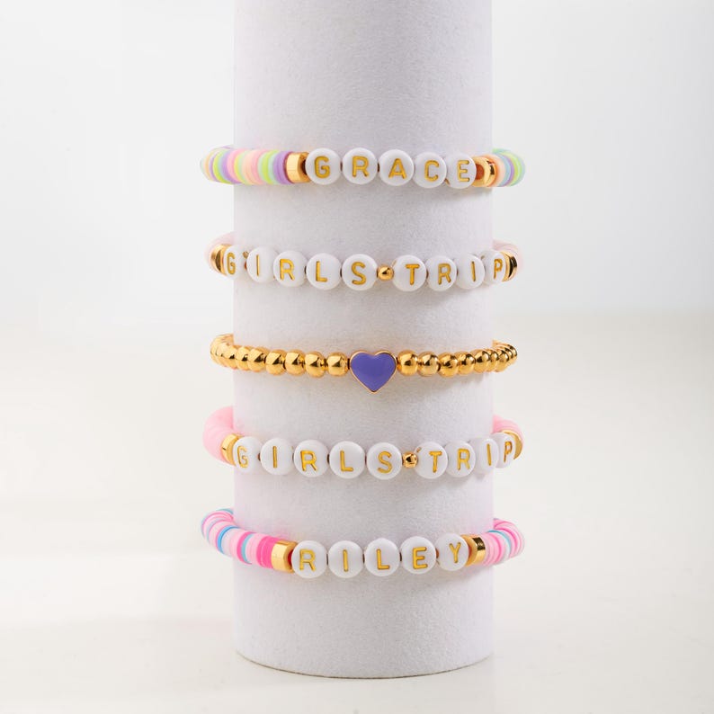 May include: Five colourful beaded bracelets with gold lettering. The bracelets spell out the names "GRACE", "GIRLS TRIP", "RILEY", and "GIRLS TRIP" twice. One bracelet has a purple heart charm.
