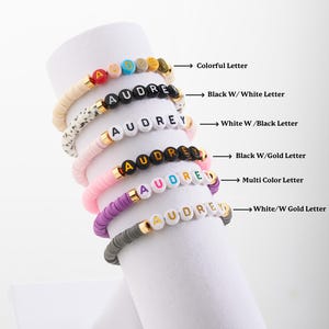 May include: A stack of seven bracelets with the name "AUDREY" spelled out in different coloured beads. The bracelets feature beads in white, black, pink, purple, grey, and multi-coloured beads with gold letters.