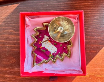 May include: A decorative, leaf-shaped trinket dish with a gold rim and a magenta interior. The dish features a white cow illustration and is topped with a small, gold-colored bowl. The dish is presented in a red box with pink tissue paper.