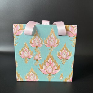 May include: A light blue gift bag with pink and gold lotus flower designs and pink ribbon handles.