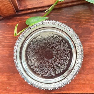 May include: A silver-colored, round serving tray with an ornate floral and geometric pattern in the center. The tray has a decorative, beaded edge. A green plant with a few leaves is in the upper left corner.