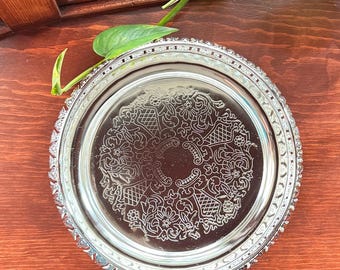 May include: A silver-colored, round serving tray with an ornate floral and geometric pattern in the center. The tray has a decorative, beaded edge. A green plant with a few leaves is in the upper left corner.