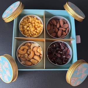 May include: A blue and gold gift box with four compartments filled with different snacks. The compartments contain almonds, dried cranberries, cashews, and puffed rice.