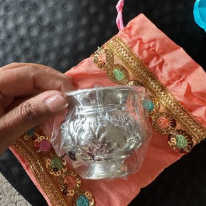 May include: A silver metal container with an ornate design is wrapped in plastic and placed inside a peach-colored fabric pouch with gold and colorful sequins.