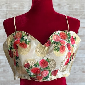 May include: A white crop top with a floral print of red roses and green leaves on a gold background. The top has a sweetheart neckline and thin straps.