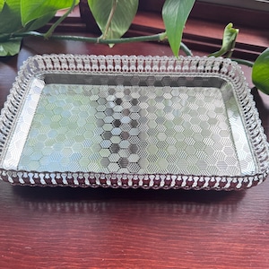 May include: A rectangular silver-colored tray with a patterned surface and a decorative, scalloped edge. The tray's interior features a geometric design. The tray is made of metal and is suitable for serving or display.