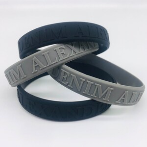 Könnte beinhalten: Drei Silikonarmbänder mit dem geprägten Text "DENIM ALEXAND". Zwei Armbänder sind schwarz und eines ist grau.