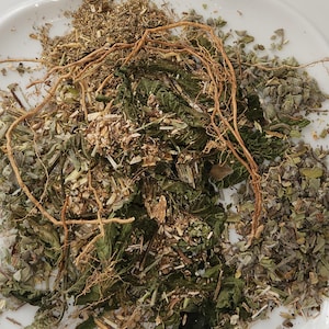 May include: A white plate filled with a selection of dried herbs and roots. The herbs are a mix of green, brown, and tan, with visible stems and leaves. The roots are a light brown colour and are intertwined among the herbs.