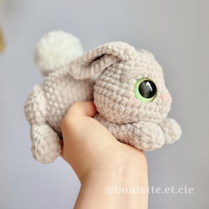 May include: A handmade, light gray crochet bunny with large, green-eyed, and a fluffy white tail. The bunny has pink accents on its cheeks and is held in a hand. The product is a soft toy.