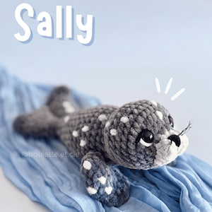 Peut inclure: Une peluche de phoque grise au crochet avec des pois blancs, posée sur un tissu bleu. Le phoque a des yeux noirs et des moustaches. Le mot "Sally" est écrit en blanc au-dessus du phoque.