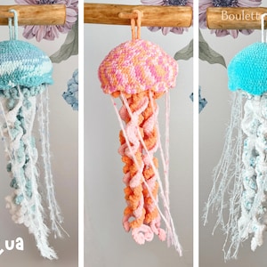 Peut inclure: Trois méduses en crochet suspendues à une branche de bois. Les méduses sont bleues, roses et orange, avec des tentacules blancs.