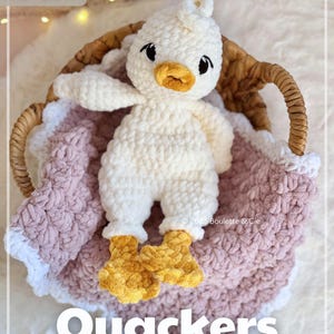 Puede incluir: Un juguete de patito blanco tejido a crochet, con pico y patas amarillas, en una cesta de mimbre sobre una manta rosa. La palabra "Quackers" se muestra en letras blancas grandes.