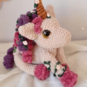 Pode incluir: Um unic&oacute;rnio de pel&uacute;cia feito &agrave; m&atilde;o com um corpo rosa claro, adornado com detalhes florais roxos, rosa e verdes. O unic&oacute;rnio tem um chifre marrom e olhos grandes e expressivos. O brinquedo de pel&uacute;cia est&aacute; sentado.