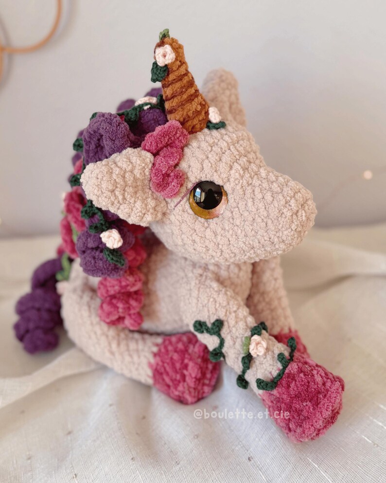 Pode incluir: Um unic&oacute;rnio de pel&uacute;cia feito &agrave; m&atilde;o com um corpo rosa claro, adornado com detalhes florais roxos, rosa e verdes. O unic&oacute;rnio tem um chifre marrom e olhos grandes e expressivos. O brinquedo de pel&uacute;cia est&aacute; sentado.