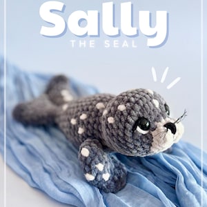 K&ouml;nnte beinhalten: Ein handgefertigtes geh&auml;keltes Seehund-Pl&uuml;schtier in Grau und Wei&szlig; mit dem Text "Sally the Seal". Der Seehund hat wei&szlig;e Flecken, schwarze Augen und eine wei&szlig;e Schnauze. Er liegt auf blauem Stoff.