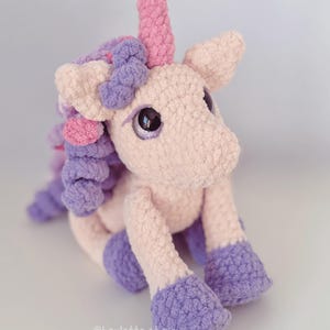 Pode incluir: Um unic&oacute;rnio de pel&uacute;cia feito &agrave; m&atilde;o em cores pastel suaves. O unic&oacute;rnio apresenta um corpo rosa, cascos e crina roxos e um chifre rosa. Olhos grandes e reflexivos d&atilde;o ao brinquedo uma apar&ecirc;ncia caprichosa. O brinquedo &eacute; feito de um material texturizado e difuso.