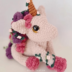 Peut inclure: Une peluche licorne rose pâle avec une corne marron et de grands yeux dorés. La crinière et la queue sont ornées de fleurs violettes, roses et vertes. Les pattes de la licorne sont décorées de guirlandes florales roses et vertes.