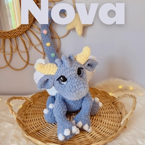 Peut inclure: Une peluche dragon crochetée bleue avec des cornes jaunes, des ailes blanches et une longue queue décorée de petits points colorés. Le texte Nova et The Sky Dragon est visible, ainsi que le texte designed by Boulette & Cie.