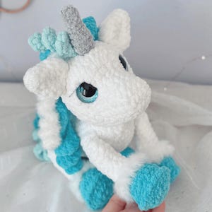 Pode incluir: Um brinquedo de pel&uacute;cia de unic&oacute;rnio feito &agrave; m&atilde;o em branco e turquesa. O unic&oacute;rnio tem um chifre prateado, olhos azuis e detalhes turquesa na crina, cauda e cascos. O brinquedo &eacute; feito de material macio e texturizado.
