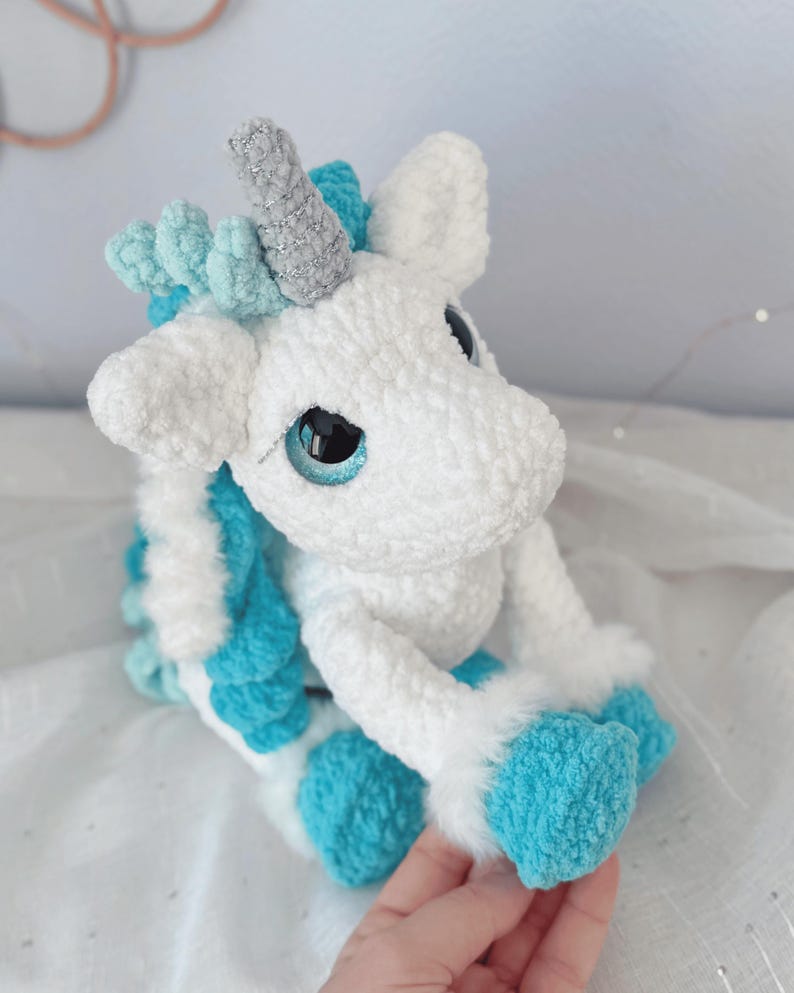Pode incluir: Um brinquedo de pel&uacute;cia de unic&oacute;rnio feito &agrave; m&atilde;o em branco e turquesa. O unic&oacute;rnio tem um chifre prateado, olhos azuis e detalhes turquesa na crina, cauda e cascos. O brinquedo &eacute; feito de material macio e texturizado.