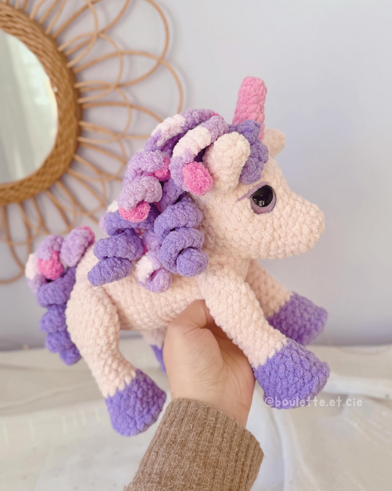 Pode incluir: Um brinquedo de unic&oacute;rnio de pel&uacute;cia feito &agrave; m&atilde;o em cores pastel suaves. O unic&oacute;rnio tem um chifre rosa, uma crina roxa e rosa e cascos roxos. O corpo &eacute; rosa claro. O brinquedo &eacute; segurado em uma m&atilde;o.