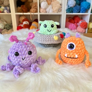 Puede incluir: Tres juguetes de peluche de crochet: un pulpo morado, un monstruo naranja con un ojo y un extraterrestre verde en una nave espacial gris.
