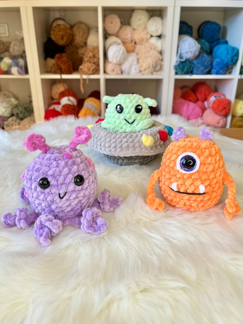 Puede incluir: Tres juguetes de peluche de crochet: un pulpo morado, un monstruo naranja con un ojo y un extraterrestre verde en una nave espacial gris.