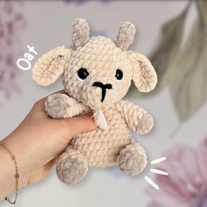 May include: A crocheted goat plush toy with a beige color and black eyes. The toy is sitting in a person's hand. The text "Oat" is written in white on a pink background.