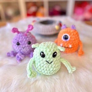 Puede incluir: Tres juguetes de peluche de crochet con forma de monstruos. El monstruo de la izquierda es morado con antenas, el monstruo del medio es verde con ojos grandes y el monstruo de la derecha es naranja con un ojo grande.