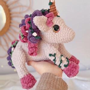 Pode incluir: Unic&oacute;rnio de croch&ecirc; feito &agrave; m&atilde;o em rosa claro, com um chifre marrom e olhos grandes. Possui detalhes roxos e rosa na crina, cauda e pernas, com enfeites florais. O unic&oacute;rnio &eacute; segurado em uma m&atilde;o, mostrando sua qualidade artesanal.