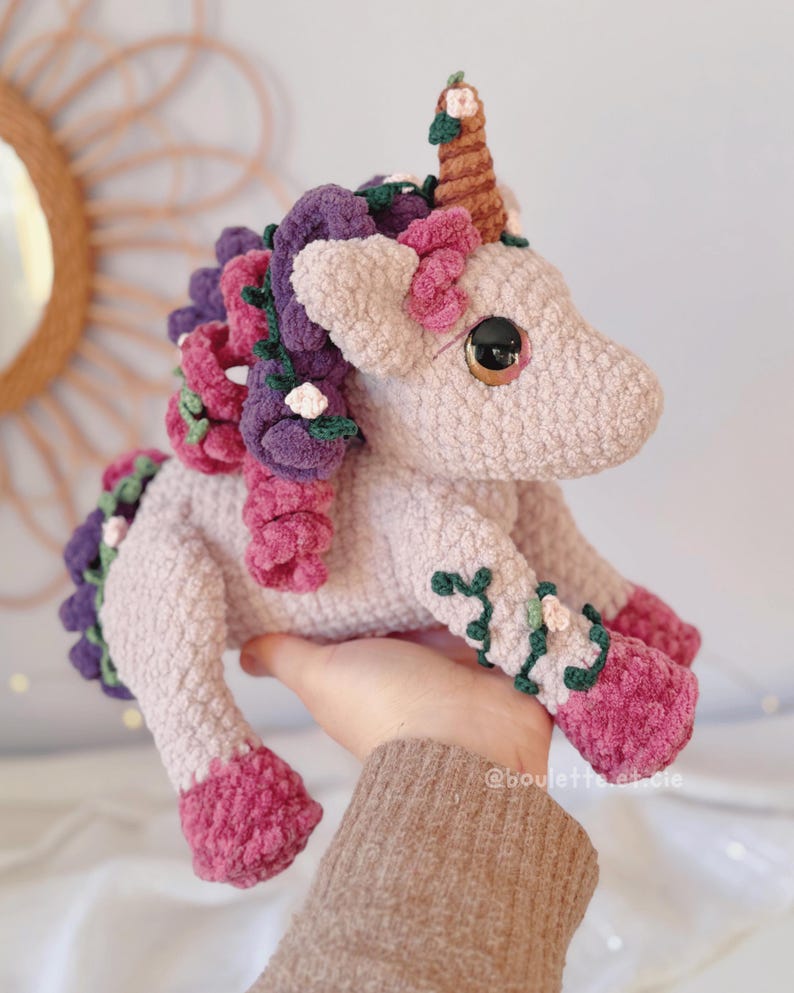 Pode incluir: Unic&oacute;rnio de croch&ecirc; feito &agrave; m&atilde;o em rosa claro, com um chifre marrom e olhos grandes. Possui detalhes roxos e rosa na crina, cauda e pernas, com enfeites florais. O unic&oacute;rnio &eacute; segurado em uma m&atilde;o, mostrando sua qualidade artesanal.
