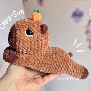 Peut inclure: Un jouet en peluche capybara en crochet marron avec une petite carotte orange sur la tête. Le jouet est tenu par une main. Le texte "Capy-Sun" est écrit en blanc sur un fond blanc.