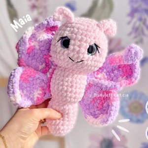 Op de afbeelding: Een roze en paarse gehaakte vlinder speelgoed met grote ogen en de naam "Maia" erboven geschreven. Het speelgoed wordt in een hand gehouden en de achtergrond is wazig.