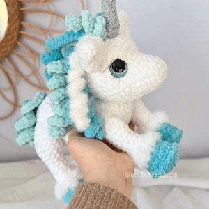 Pode incluir: Um brinquedo de pel&uacute;cia de unic&oacute;rnio de croch&ecirc; branco e azul feito &agrave; m&atilde;o. O unic&oacute;rnio tem um chifre prateado, crina e cauda azuis e azul claro e olhos azuis grandes. O brinquedo de pel&uacute;cia &eacute; segurado em uma m&atilde;o.
