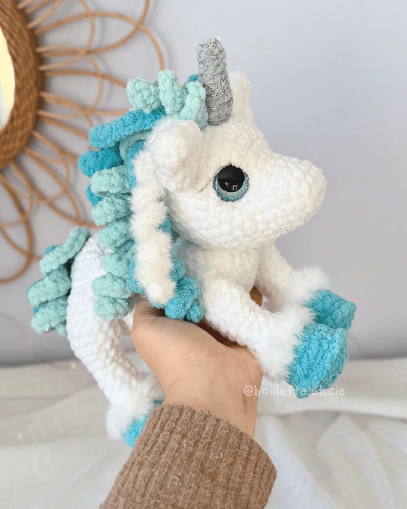 Pode incluir: Um brinquedo de pel&uacute;cia de unic&oacute;rnio de croch&ecirc; branco e azul feito &agrave; m&atilde;o. O unic&oacute;rnio tem um chifre prateado, crina e cauda azuis e azul claro e olhos azuis grandes. O brinquedo de pel&uacute;cia &eacute; segurado em uma m&atilde;o.