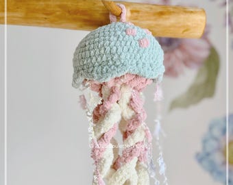 May include: A handcrafted crochet jellyfish mobile in pastel shades. The jellyfish has a pale blue cap with pink spots and long, braided tentacles in white and pink. The word "Acqua" is displayed above the jellyfish.