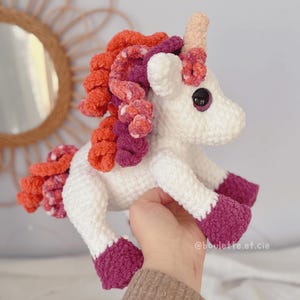 Pode incluir: Um unic&oacute;rnio branco feito &agrave; m&atilde;o em croch&ecirc;, com um chifre rosa, olhos pretos e uma crina colorida de fios laranja, rosa e roxo. Os cascos do unic&oacute;rnio s&atilde;o de cor roxa escura. A imagem &eacute; de um bicho de pel&uacute;cia.