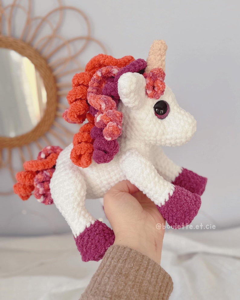 Pode incluir: Um unic&oacute;rnio branco feito &agrave; m&atilde;o em croch&ecirc;, com um chifre rosa, olhos pretos e uma crina colorida de fios laranja, rosa e roxo. Os cascos do unic&oacute;rnio s&atilde;o de cor roxa escura. A imagem &eacute; de um bicho de pel&uacute;cia.