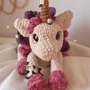 Pode incluir: Um unic&oacute;rnio de croch&ecirc; feito &agrave; m&atilde;o em rosa claro, com um chifre marrom e olhos grandes. &Eacute; adornado com detalhes florais rosa, roxos e verdes, com cascos rosa combinando. Este brinquedo de pel&uacute;cia &eacute; uma cria&ccedil;&atilde;o charmosa e caprichosa.