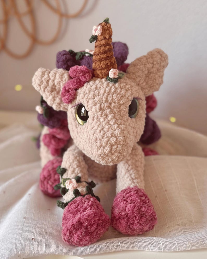 Pode incluir: Um unic&oacute;rnio de croch&ecirc; feito &agrave; m&atilde;o em rosa claro, com um chifre marrom e olhos grandes. &Eacute; adornado com detalhes florais rosa, roxos e verdes, com cascos rosa combinando. Este brinquedo de pel&uacute;cia &eacute; uma cria&ccedil;&atilde;o charmosa e caprichosa.