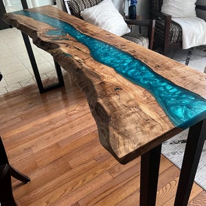May include: A live edge wooden table with a vibrant blue epoxy resin river running through the center. The table is supported by black metal legs. The wood has a natural, light brown colour.