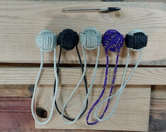 May include: Five decorative rope balls in various colors, including gray, black, and purple, each with a loop of rope attached. The balls are arranged on a wooden surface, with a pen visible in the background.
