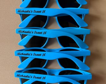May include: Five pairs of blue plastic sunglasses with black lenses. Each pair has the text "Michaela's Sweet 16" printed on the arms.