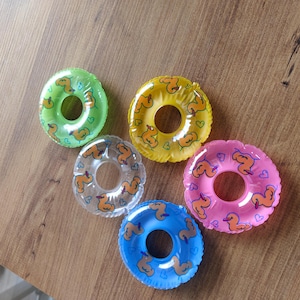 Op de afbeelding: Vijf kleine, ronde, opblaasbare drankhouders in groen, geel, transparant, roze en blauw. Elke drijver is voorzien van een herhalend patroon van oranje badeendjes en kleine blauwe hartjes, gedrukt op het glanzende, doorschijnende plastic oppervlak.