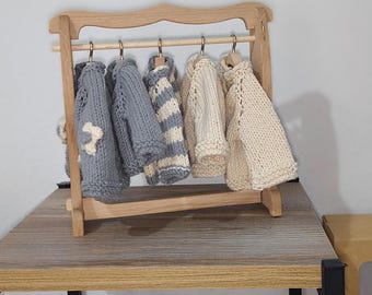 May include: A wooden clothes rack with five hangers holding knitted sweaters in shades of gray and beige. The rack is on a wooden shelf.