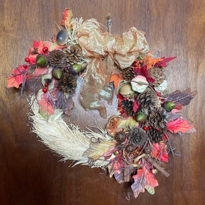 Może przedstawiać: Jesienny wieniec z podstawą ze słomy, ozdobiony sztucznymi liśćmi w odcieniach czerwieni, pomarańczy i brązu. Zawiera szyszki, żołędzie i dużą złotą wstążkę. Idealny do jesiennej dekoracji domu, około 30 cm średnicy.