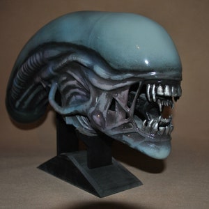 Può includere: Una scultura dettagliata di una testa aliena dipinta a mano con uno schema di colori blu e grigio. La scultura è montata su un piedistallo nero e ha un'espressione minacciosa con denti affilati.