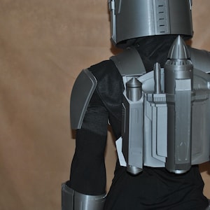 Puede incluir: Un disfraz gris al estilo mandaloriano, que incluye un casco, una armadura para los hombros y un jetpack. El jetpack presenta múltiples componentes cilíndricos y rectangulares. El disfraz se usa sobre un atuendo negro.